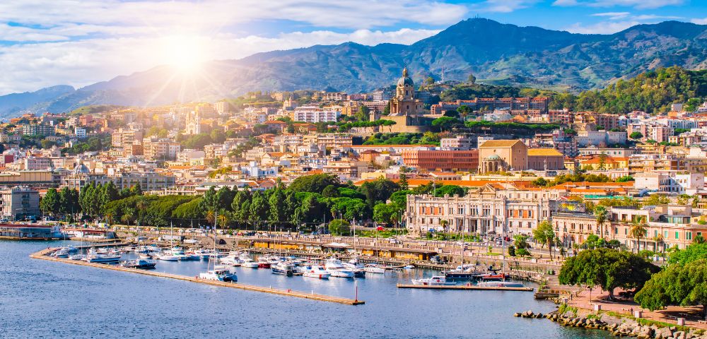 Panoramic view of a coastal city with a marina full of boats and buildings climbing a hillside under a sunny, blue sky with scattered clouds.