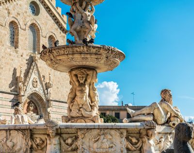 Ornate marble fountain with detailed sculptures of mythological figures and cherubs, set against a backdrop of a stone church facade under a bright blue sky with scattered clouds.