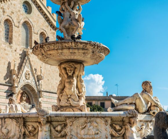 Ornate marble fountain with detailed sculptures of mythological figures and cherubs, set against a backdrop of a stone church facade under a bright blue sky with scattered clouds.