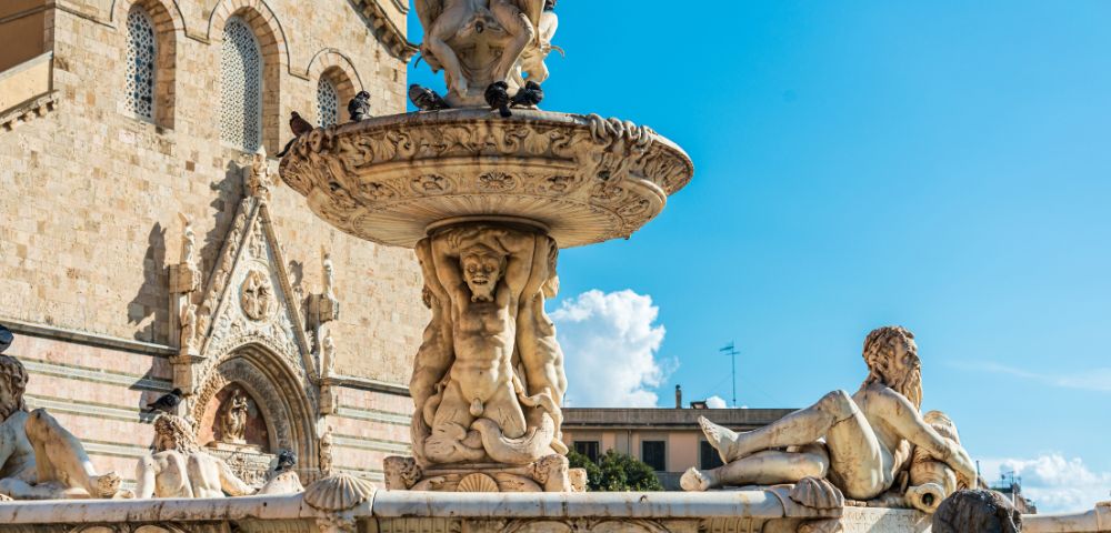 Ornate marble fountain with detailed sculptures of mythological figures and cherubs, set against a backdrop of a stone church facade under a bright blue sky with scattered clouds.