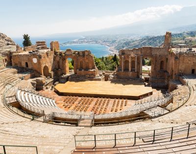 Ancient Greco-Roman theatre with stone seating tiers and a wooden stage, set against a backdrop of a coastal town and a distant mountain.