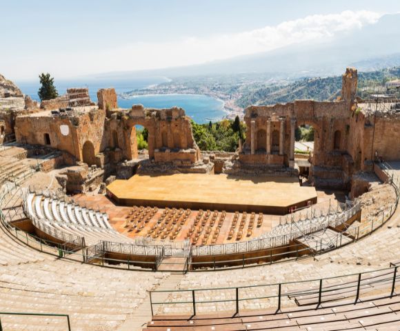 Ancient Greco-Roman theatre with stone seating tiers and a wooden stage, set against a backdrop of a coastal town and a distant mountain.