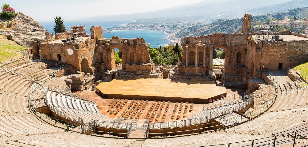 Ancient Greco-Roman theatre with stone seating tiers and a wooden stage, set against a backdrop of a coastal town and a distant mountain.
