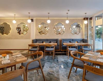 A modern restaurant interior with wooden tables and chairs, decorated with wall mirrors and soft lighting. The space has a cozy atmosphere, featuring a mix of gray and marble surfaces, and is illuminated by pendant lights.