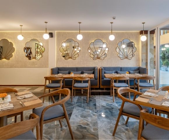 A modern restaurant interior with wooden tables and chairs, decorated with wall mirrors and soft lighting. The space has a cozy atmosphere, featuring a mix of gray and marble surfaces, and is illuminated by pendant lights.