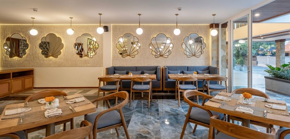 A modern restaurant interior with wooden tables and chairs, decorated with wall mirrors and soft lighting. The space has a cozy atmosphere, featuring a mix of gray and marble surfaces, and is illuminated by pendant lights.