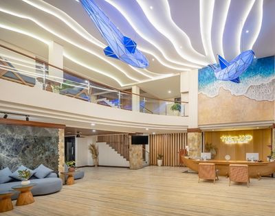 A modern hotel lobby with a spacious design, featuring a reception desk, comfortable seating, and decorative ceiling lights, as well as artistic wall murals and sculptures resembling ocean waves and marine life.