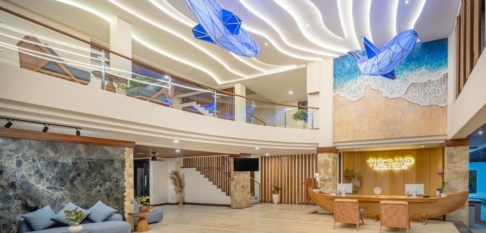 A modern hotel lobby with a spacious design, featuring a reception desk, comfortable seating, and decorative ceiling lights, as well as artistic wall murals and sculptures resembling ocean waves and marine life.