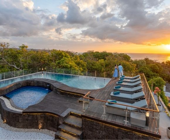 A luxurious infinity pool overlooking a sunset with two people standing by the edge, surrounded by lush greenery.