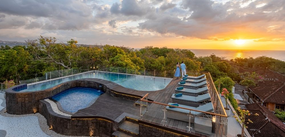 A luxurious infinity pool overlooking a sunset with two people standing by the edge, surrounded by lush greenery.