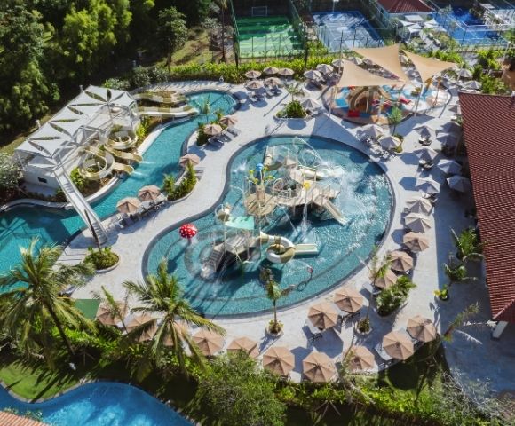 Aerial view of a large, curved swimming pool surrounded by palm trees, lounge chairs, and umbrellas, with water slides and play structures.