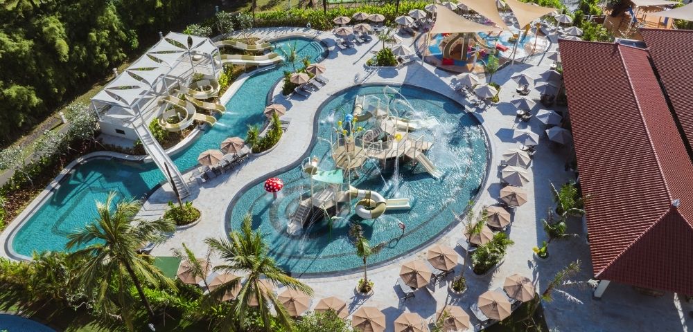 Aerial view of a large, curved swimming pool surrounded by palm trees, lounge chairs, and umbrellas, with water slides and play structures.
