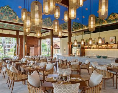 A beautifully designed restaurant with wooden pillars, modern lighting, and a tropical ambiance, featuring tables and chairs set for dining.