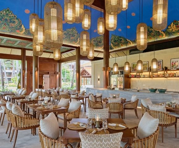 A beautifully designed restaurant with wooden pillars, modern lighting, and a tropical ambiance, featuring tables and chairs set for dining.