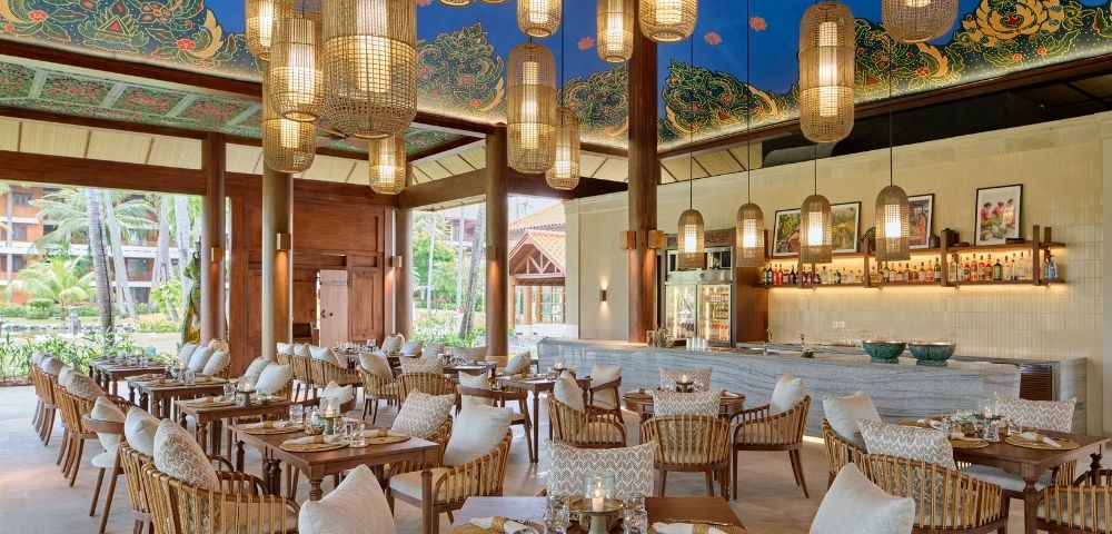 A beautifully designed restaurant with wooden pillars, modern lighting, and a tropical ambiance, featuring tables and chairs set for dining.
