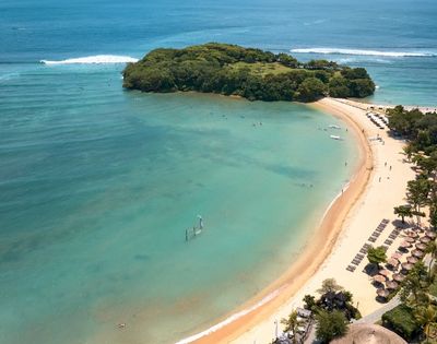 Aerial view of a beach with clear water, an island in the distance, and people enjoying the shoreline.
