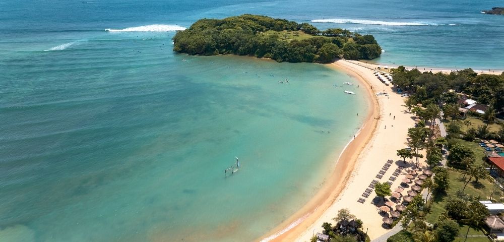 Aerial view of a beach with clear water, an island in the distance, and people enjoying the shoreline.