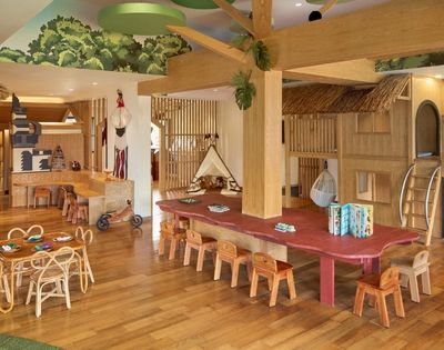 A spacious wooden playroom with various play structures, a large table surrounded by small chairs, and wooden decor. The ceiling features greenery, and there are toys and activity areas for children.