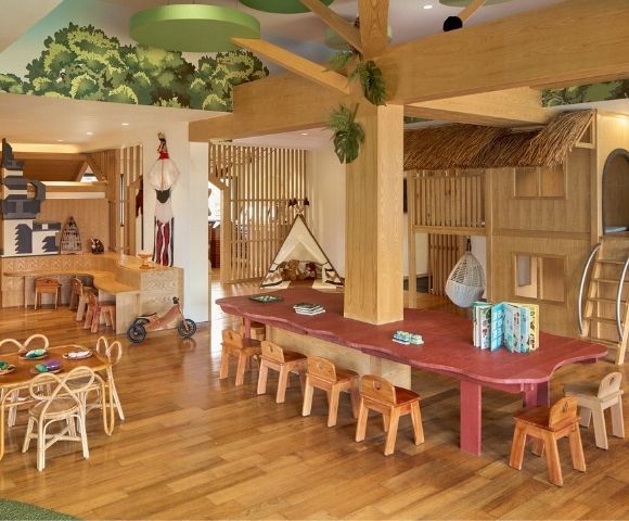 A spacious wooden playroom with various play structures, a large table surrounded by small chairs, and wooden decor. The ceiling features greenery, and there are toys and activity areas for children.