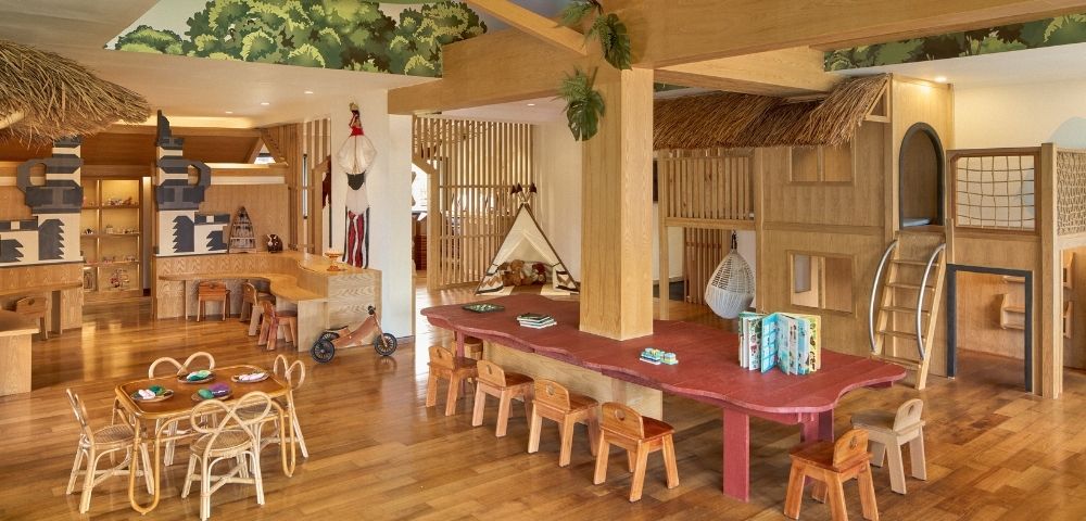 A spacious wooden playroom with various play structures, a large table surrounded by small chairs, and wooden decor. The ceiling features greenery, and there are toys and activity areas for children.