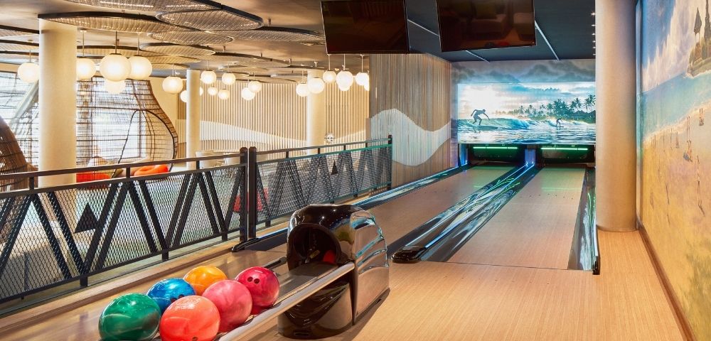A modern bowling alley featuring multiple lanes, colorful bowling balls on a rack, and decorative murals on the walls.