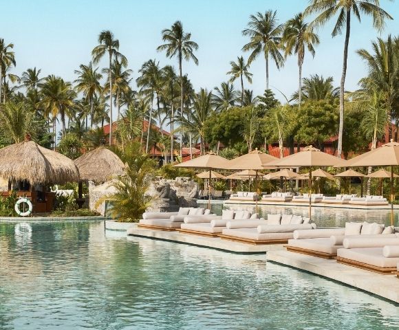 A serene resort pool area with palm trees, thatched-roof cabanas, and lounge chairs under umbrellas.