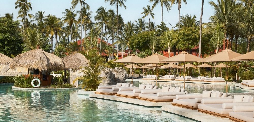 A serene resort pool area with palm trees, thatched-roof cabanas, and lounge chairs under umbrellas.