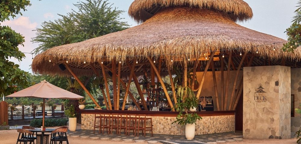 A tropical outdoor bar with a thatched roof, surrounded by trees and decorative lights.