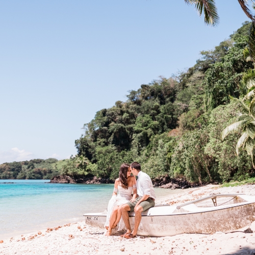 Couple sits affectionately on a small boat beside a white tropical beach, with turquoise water, lush jungle, and a peaceful romantic mood.