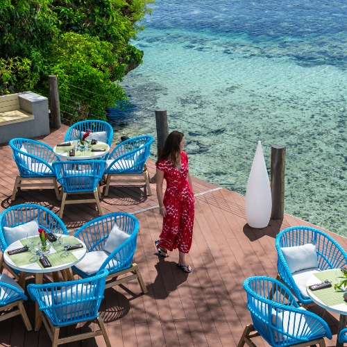 Oceanfront restaurant deck with turquoise chairs and tables, a woman in a red dress, lush greenery, and clear blue water beyond.