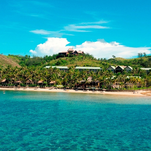 Turquoise tropical sea beside a sandy beach, palm-lined resort, and green hillside villas under bright blue skies; serene vacation atmosphere.