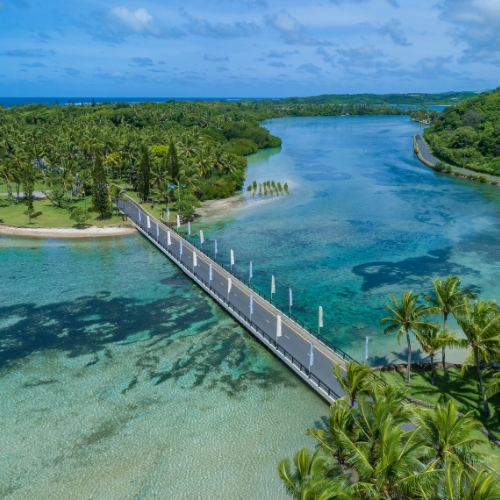 Aerial view of a bridge crossing turquoise lagoon waters, surrounded by lush palm trees, tropical shoreline, and a calm blue horizon.