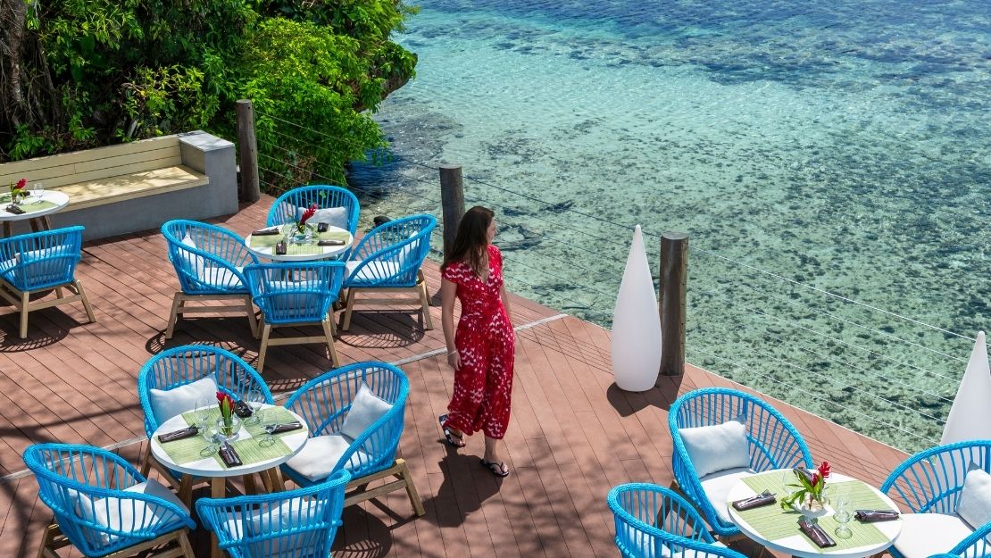 Oceanfront restaurant deck with turquoise chairs and tables, a woman in a red dress, lush greenery, and clear blue water beyond.