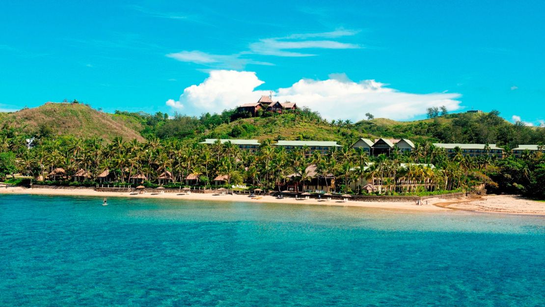 Turquoise tropical sea beside a sandy beach, palm-lined resort, and green hillside villas under bright blue skies; serene vacation atmosphere.