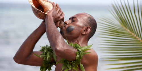 A man blows into a large conch shell beside the ocean, with palm fronds behind him, creating a festive island welcome.