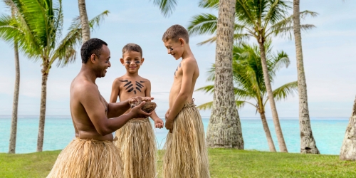Three smiling people in grass skirts gather among palm trees by a turquoise beach.