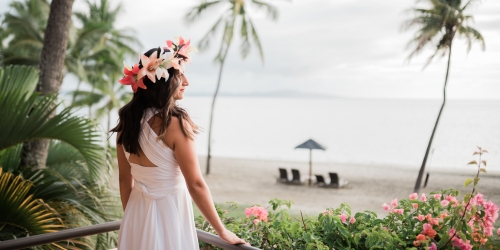 Woman in a flowing white dress and tropical flower crown gazes at a beach with palm trees, ocean, cabana, and pink flowers.