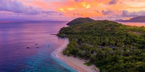 Aerial view of a tropical island coastline at sunset, with turquoise water, sandy coves, lush green hills, and vivid pink-orange clouds.
