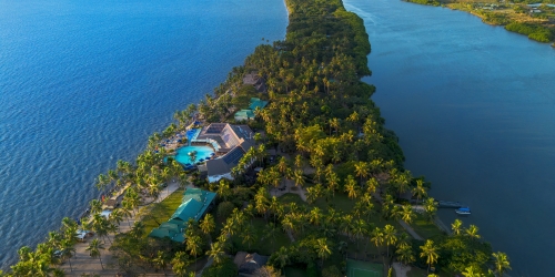 Aerial view of a lush tropical resort on a narrow peninsula between blue waters, with palm trees, pools, villas, and a serene getaway mood.