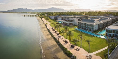 Aerial view of a serene lakeside resort with sandy beach, palm-lined walkway, turquoise pools, modern buildings, and distant mountains.