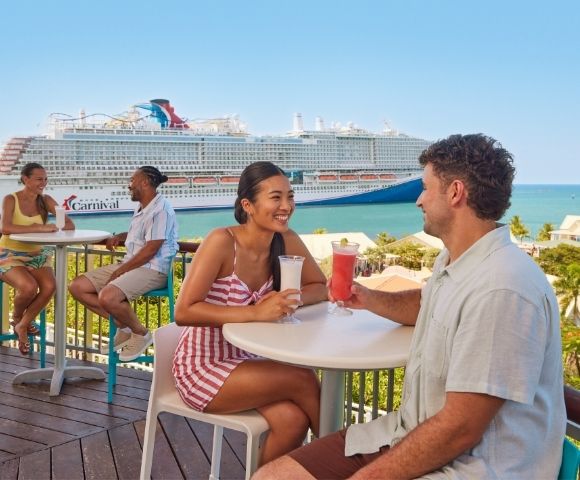 Guests enjoying drinks and conversation on an outdoor cruise ship deck overlooking the ocean.