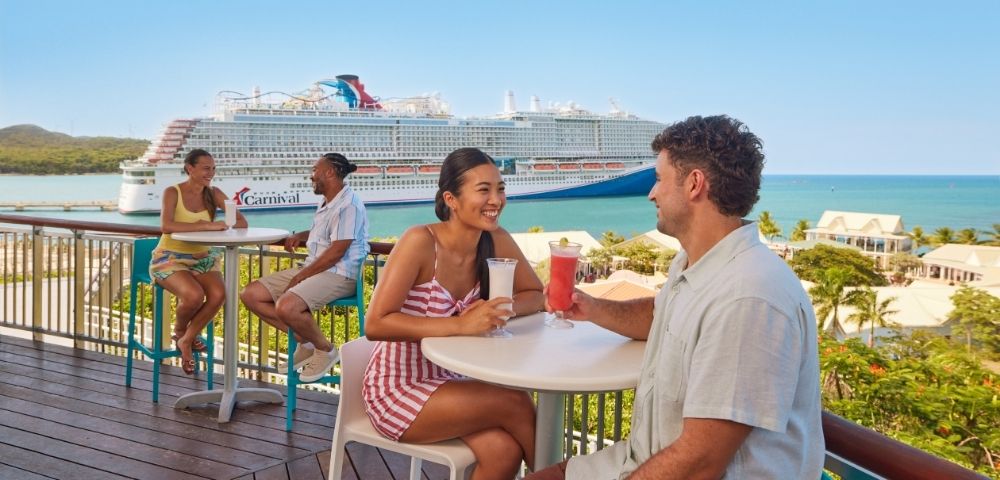 Guests enjoying drinks and conversation on an outdoor cruise ship deck overlooking the ocean.