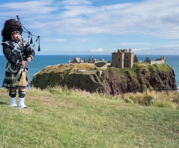 A bagpiper in traditional Scottish attire stands on a grassy cliff overlooking the sea and the stone ruins of Dunnottar Castle under a blue sky.