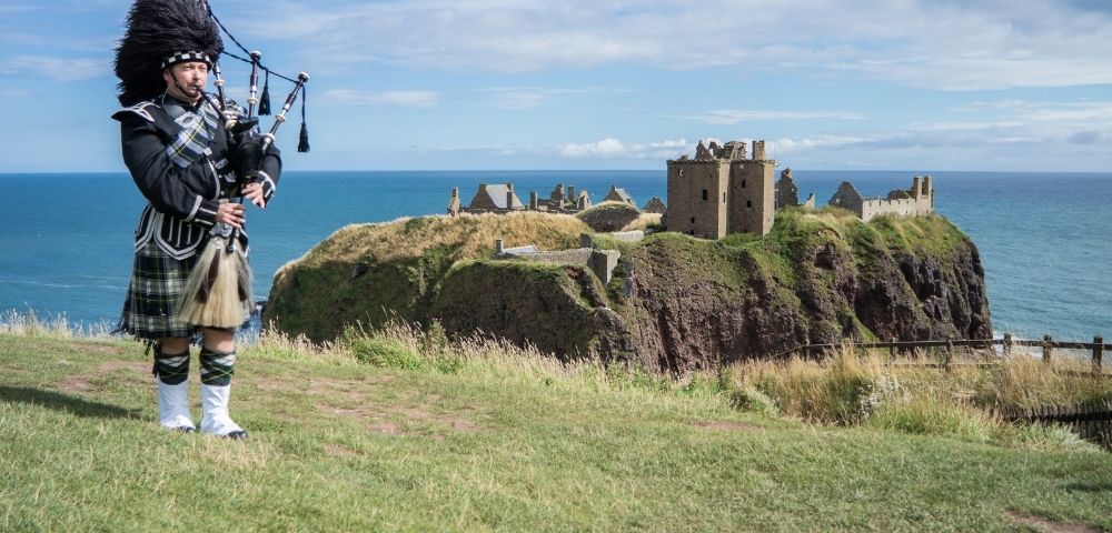 A bagpiper in traditional Scottish attire stands on a grassy cliff overlooking the sea and the stone ruins of Dunnottar Castle under a blue sky.