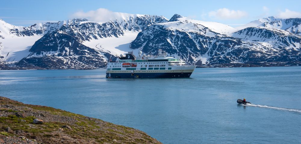 Expedition cruise ship navigates calm blue water with a rugged, snow-capped mountain range in the background. A small inflatable boat speeds across the water in the foreground.