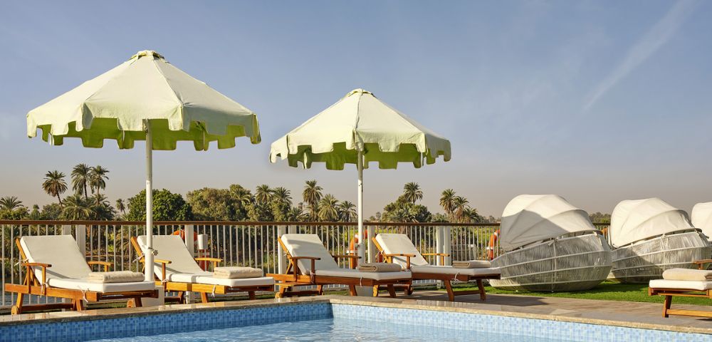 A resort poolside with light green scalloped umbrellas, wooden lounge chairs with white cushions, and unique white-canopied pod-shaped loungers, overlooking a palm-filled landscape under a bright blue sky.