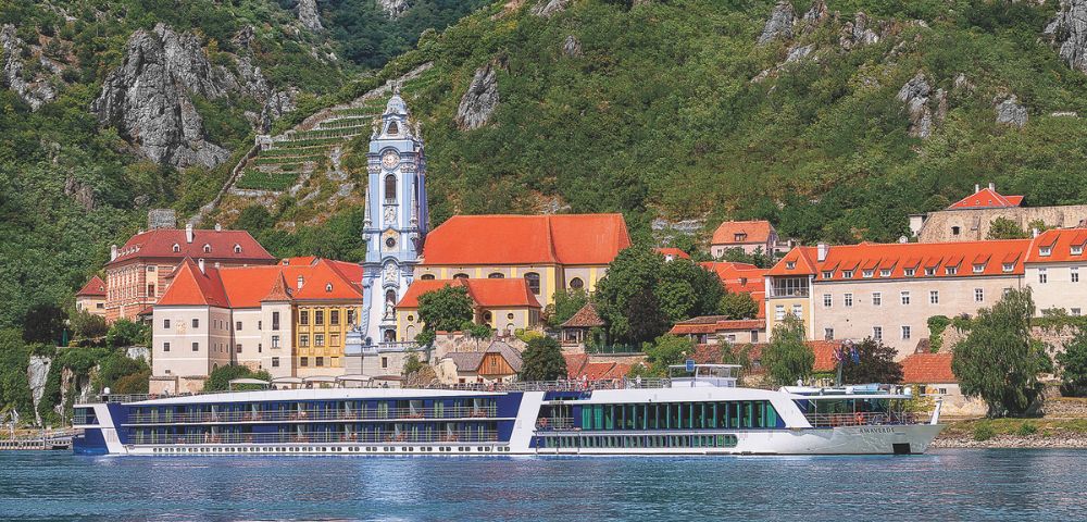 A modern, multi-deck river cruise ship sails on a wide river in front of a picturesque European town built on a hillside with steep, tree-covered mountains rising behind it. The town features traditional architecture with orange-tiled roofs, a distinctive blue and white church steeple, and historic stone buildings.
