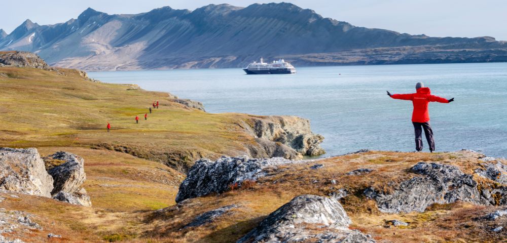A person in a red jacket stands with arms outstretched on a rocky, grassy cliff overlooking a calm bay with a large cruise ship in the distance and a line of people walking along the shore below.