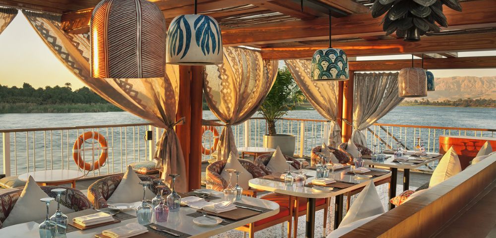 Elegant outdoor dining area on a river cruise ship deck, set with tables and chairs, illuminated by decorative pendant lights, overlooking a tranquil river and distant hills at sunset.