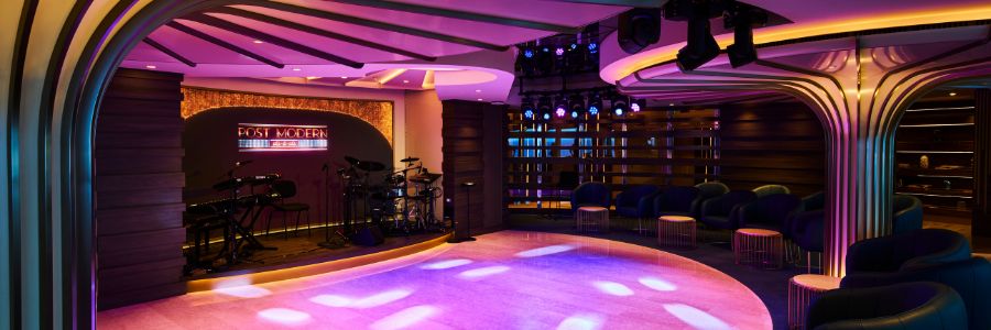 A modern lounge with a stage, DJ booth, and seating area, illuminated by purple and pink lights.